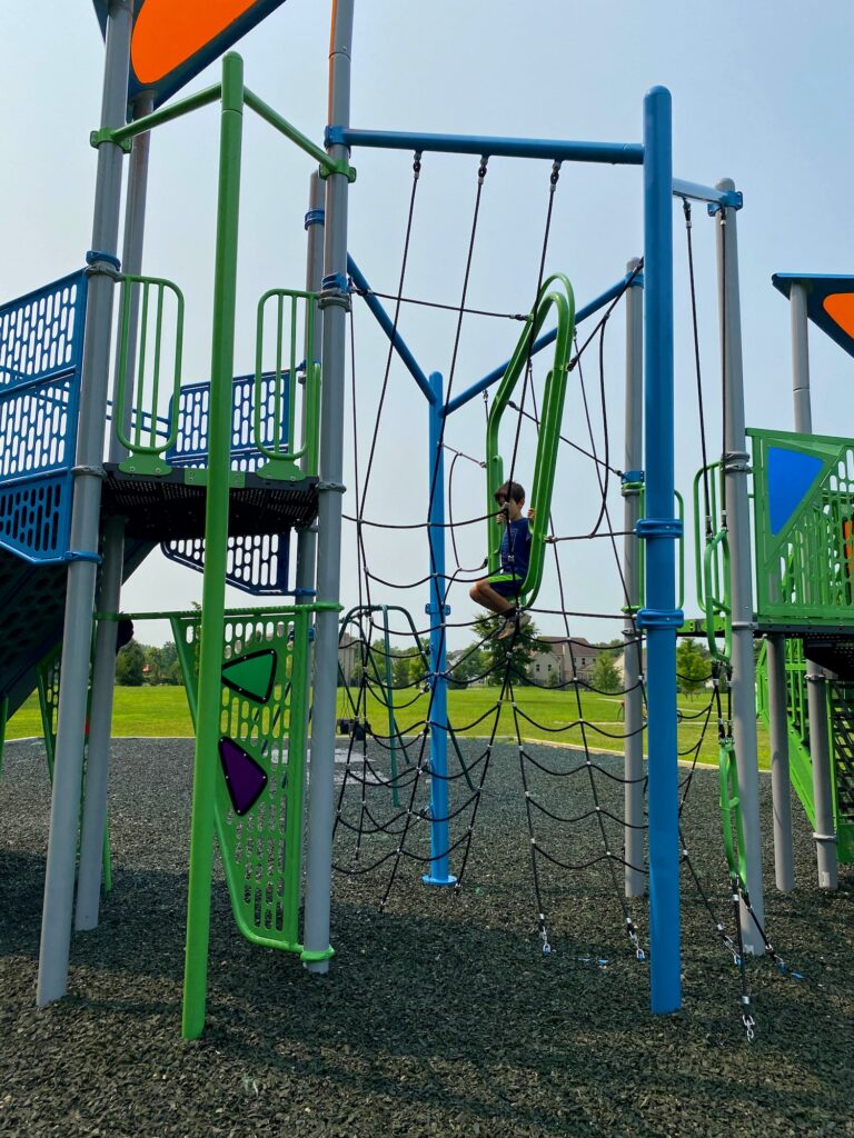 A boy sitting on a seat in the middle of a rope climber at the park.