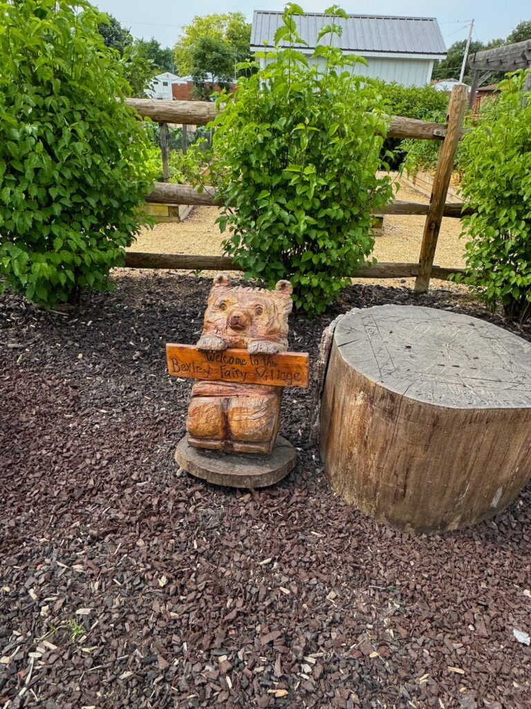 A wooden bear holding a sign that says Welcome to Bexley Fairy Village.
